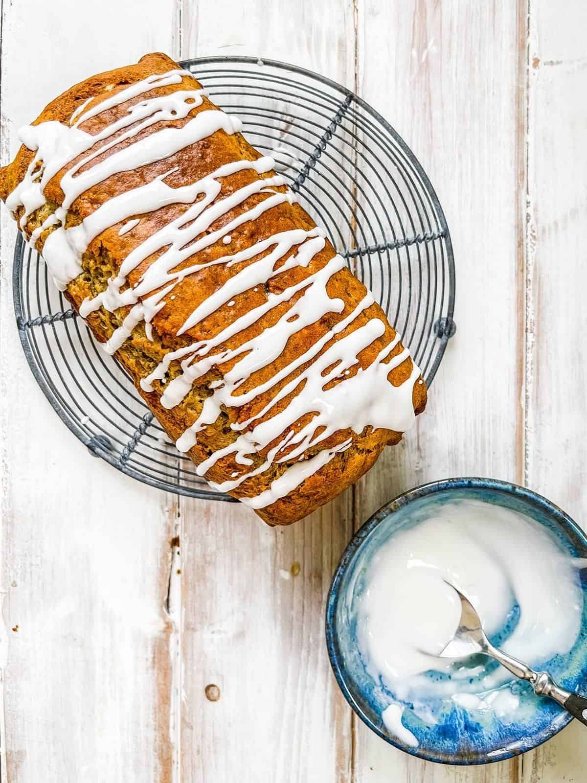 A banana loaf on a white table with a small dish of frosting to the side.