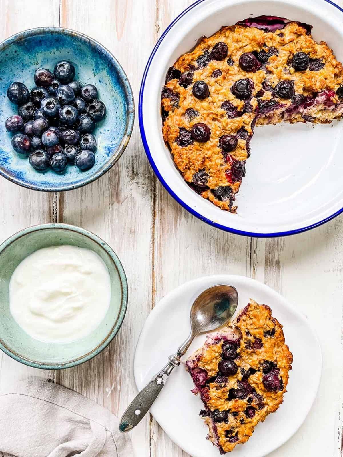 A dish of baked oatmeal on a white table with a dish of blueberries and some yogurt. 